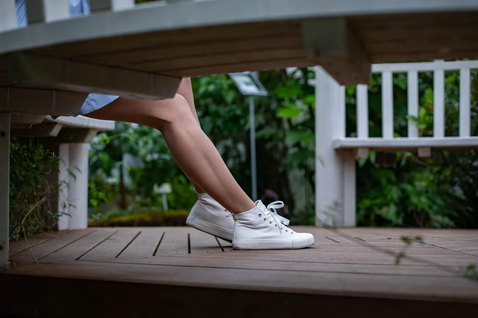 Legs of a woman in white sneakers sitting on a wooden bench in a leafy park